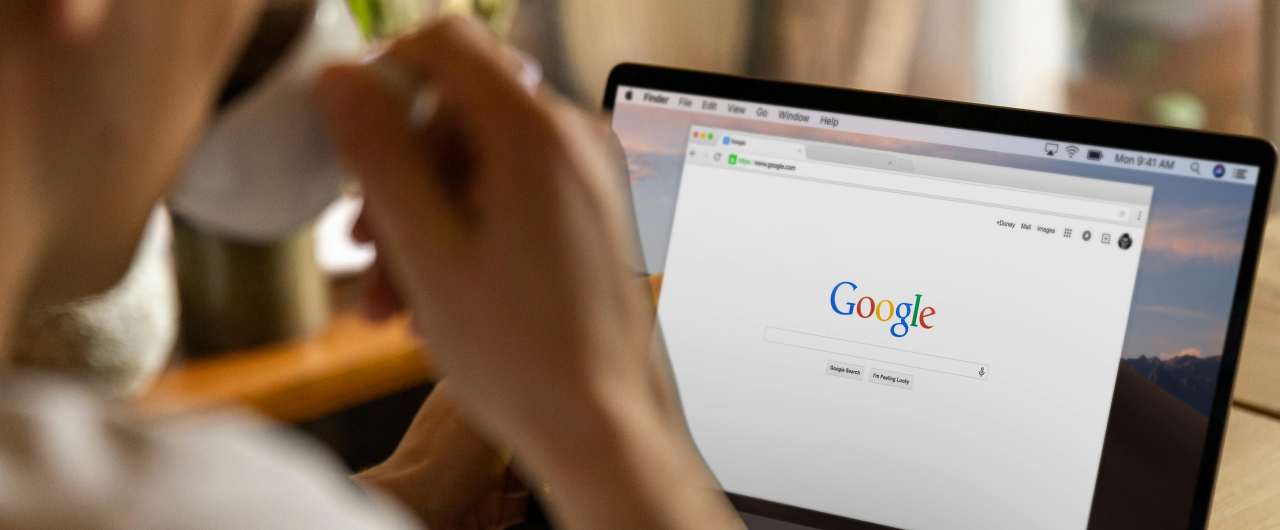 Людина тримає чашку, дивиться на ноутбук з домашньою сторінкою Google, дерев’яний стіл із рослинами на задньому плані.